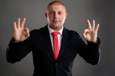 Zarif siyah takım elbiseli ve kırmızı kravatlı havalı iş adamının portresi gri arka planda çift tamam işareti gösteriyor.