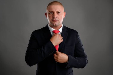 Zarif siyah takım elbiseli havalı iş adamının portresi gri arka planda izole edilmiş kırmızı kravatını düzenler.