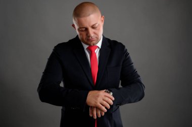 Zarif siyah takım elbiseli havalı iş adamının portresi ve kırmızı kravat gri arka planda izole edilmiş saatine bakıyor.