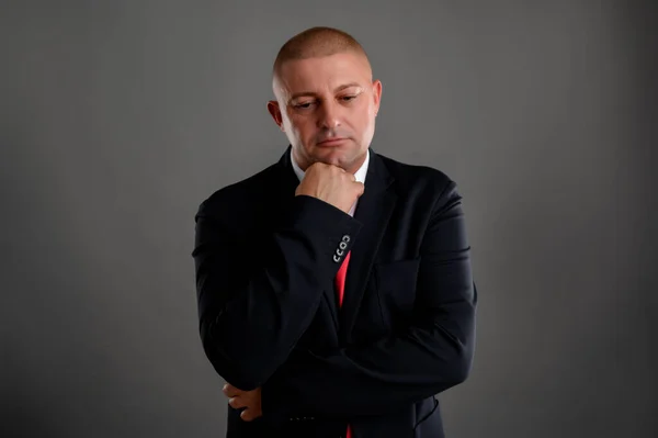 Portrait of cool businessman wearing elegant black suit and red tie thinks with hishand under his chin isolated over gray background