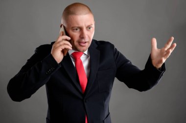 Zarif siyah takım elbiseli ve telefonda konuşan kırmızı kravatlı havalı iş adamının portresi gri arka planda gergin bir şekilde yalıtılmış bir şekilde el kol hareketi yapmaya başlıyor.