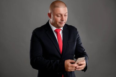 Zarif siyah takım elbise ve kırmızı kravat giyen, cep telefonu kullanan havalı iş adamının portresi gri arka planda izole edilmiş kısa mesaj yazıyor.