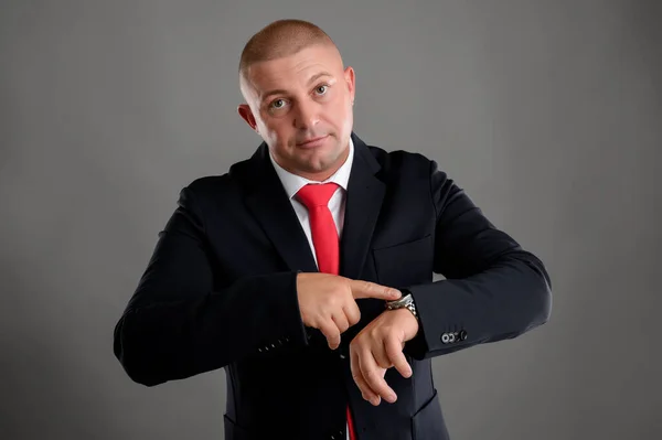 Portrait of cool businessman wearing elegant black suit and red tie showing time on his watch isolated over gray background