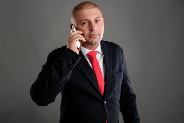 Portrait of cool businessman wearing elegant black suit and red tie talking on the phone isolated over gray background
