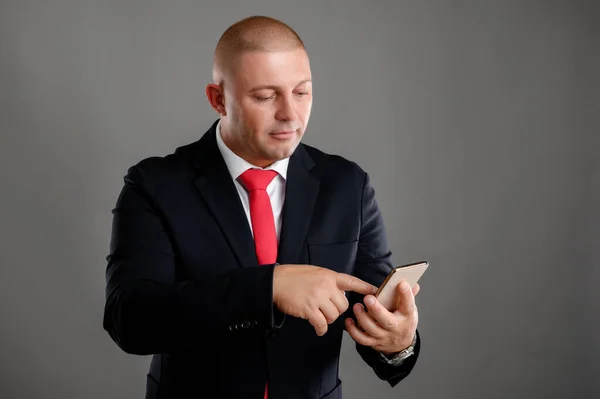 Portrait of cool businessman wearing elegant black suit and red tie using mobile phone, writing text message isolated over gray background