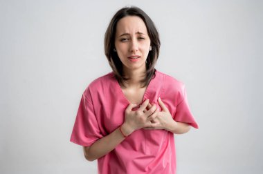 Pembe hastane kıyafetleri giyen, kahverengi saçlı, kalp ağrısı çeken ya da göğüs ağrısı çeken, beyaz, izole edilmiş arka planda kalp sorunları olan genç bir kadın hemşire ya da doktor..