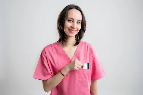 Young woman medical professional nurse or doctor dressed with pink hospital clothes, with brown hair, pull out card from her pocket posing on a white isolated backround.