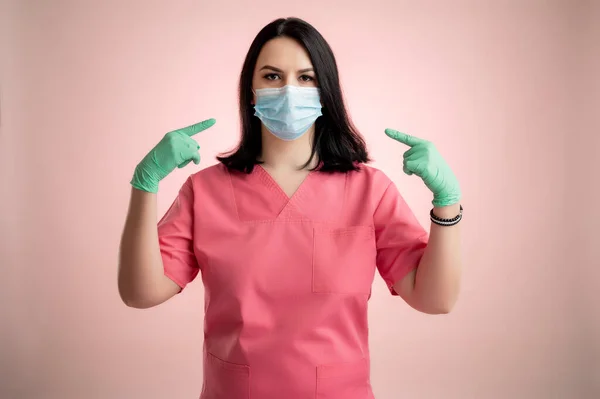 Portrait of beautiful woman doctor with stethoscope wearing pink sctubs, wears a protective mask, with her fingers pointed posing on a pink isolated backround.