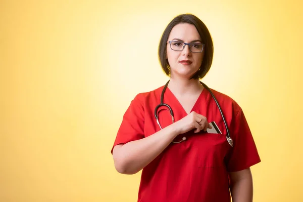Portrait of beautiful woman doctor with stethoscope wearing red scrubs, pull out card from her pocket posing on a yellow isolated background.