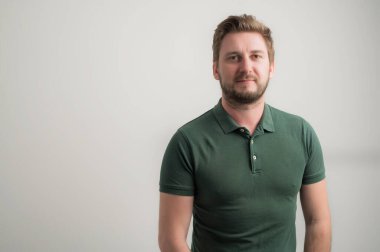 Gri arka planda izole edilmiş yeşil tişört giyen, kalın sakallı, ciddi ve şık çekici bir adamın portresi.