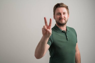 Kalın sakallı, ciddi ve çekici bir adamın portresi. Günlük yeşil tişörtler giymiş. Gri arka planda izole edilmiş barış jestleri gösteriyor.