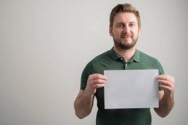 Kalın sakallı, sıradan yeşil tişörtlü, gri arka planda, fotokopi alanı reklam alanı ile izole edilmiş beyaz kağıtlı ciddi ve şık çekici bir adamın portresi.