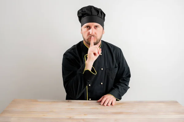 Portrait of young male chef in black uniform showing shh gesture posing on a white isolated background