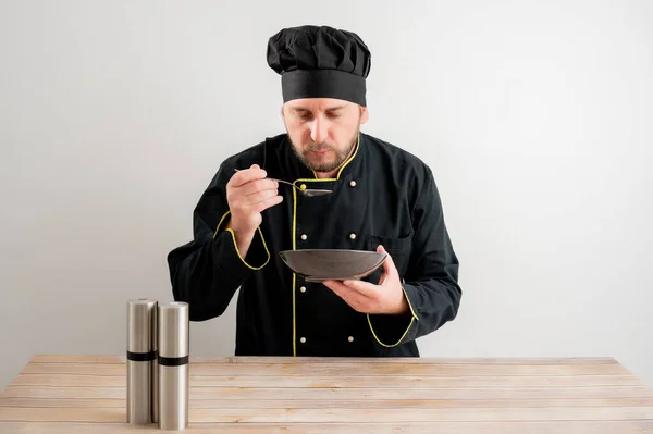 Portrait of young male chef in black uniform shows the plate in his hand posing using salt on a white isolated background