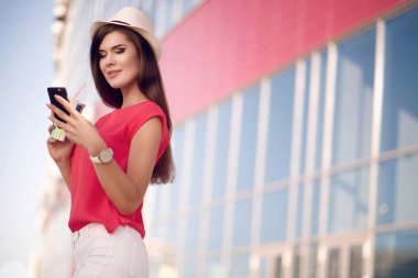 Bir moda şapka, kırmızı gömlek ve şehirde yaz aylarında açık havada kahve içme ve seyir telefon güzel uzun saçlı genç güzel kız