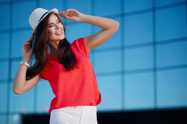 Bella ragazza felice e giovane in un cappello alla moda, camicia rossa e bei capelli lunghi che camminano nella città estiva . — Foto Stock