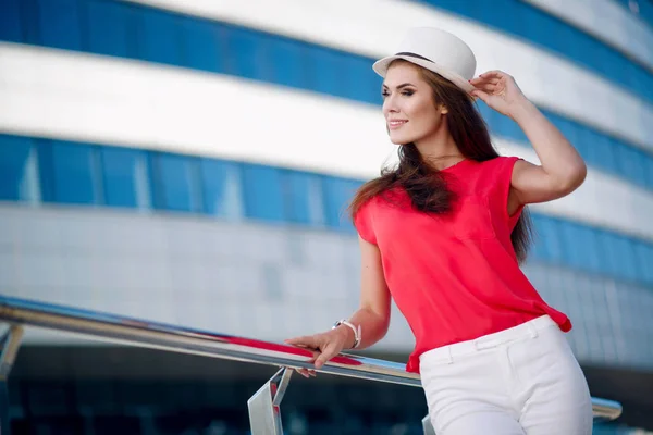 Menina bonita feliz e jovem em um chapéu na moda, camisa vermelha e cabelo longo agradável em um fundo de belo edifício azul — Fotografia de Stock