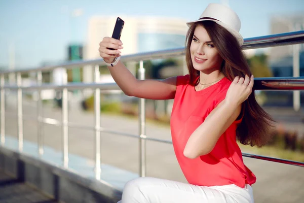 Menina bonita feliz e jovem em um chapéu na moda, camisa vermelha e belo cabelo longo torna um auto-retrato no smartphone no verão na cidade — Fotografia de Stock