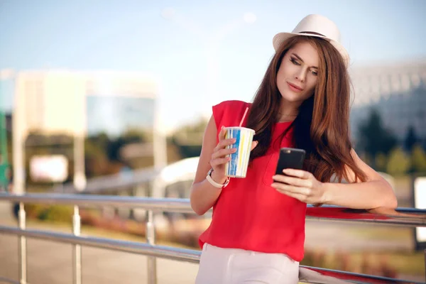 Mutlu ve genç kız güzel bir moda şapka, kırmızı gömlek ve şehirde yaz aylarında açık havada kahve içme ve seyir telefon güzel uzun saçları — Stok fotoğraf