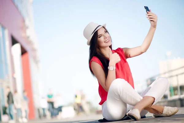 Chica hermosa feliz y joven en un sombrero de moda, camisa roja y hermoso pelo largo hace que sea un autorretrato en el teléfono inteligente en el verano en la ciudad —  Fotos de Stock