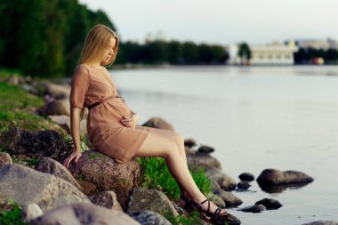 Genç güzel bir hamile kadın arka plan kentsel peyzaj yaz aylarında gölde kayaların üzerinde rahatlatıcı