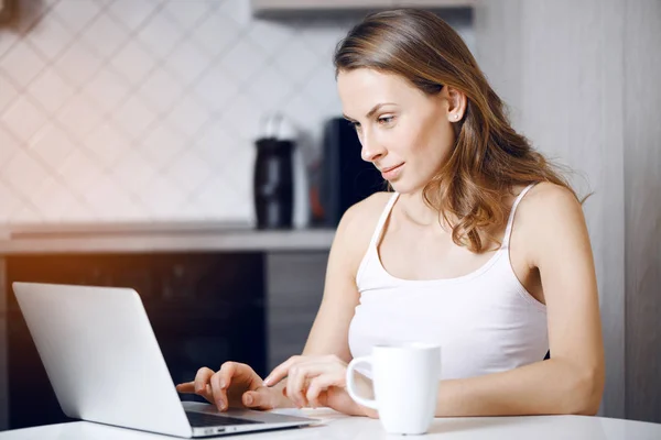 Chica se sienta a la mesa en casa y trabajando en un ordenador portátil — Foto de Stock