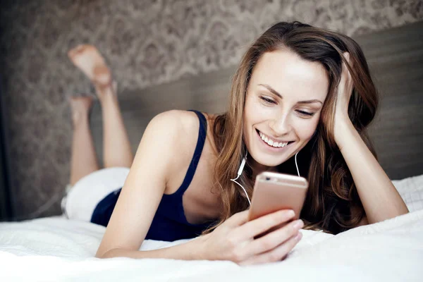 Jeune fille dans le lit dans la chambre à coucher écoutant de la musique avec des écouteurs et tenant un téléphone portable — Photo