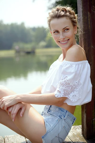 Joven chica hermosa sentada en la playa cerca del lago en el verano en pantalones cortos y sonriendo — Foto de Stock