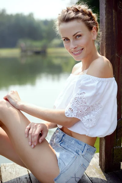 Joven chica hermosa sentada en la playa cerca del lago en el verano en pantalones cortos y sonriendo — Foto de Stock
