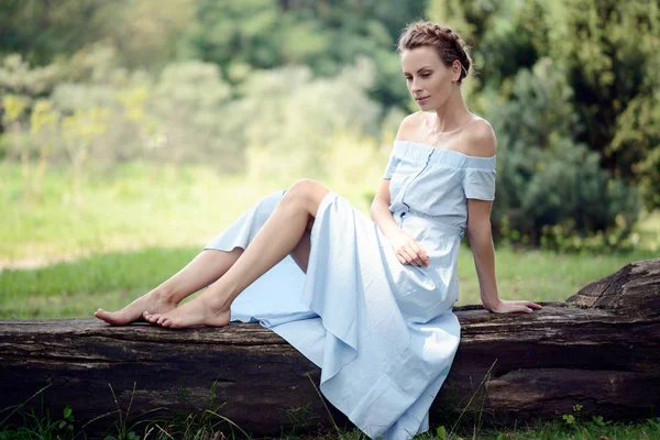 Hermosa niña delgada sentada en un tronco en el parque en el verano en un vestido azul y sonriendo —  Fotos de Stock