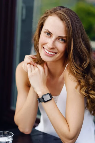 Hermosa chica sentada en un café de verano y sonriendo. Relojes inteligentes a mano — Foto de Stock