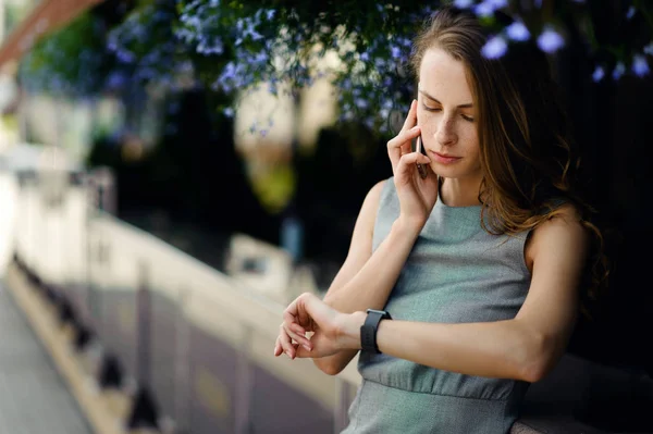 Mladá a krásná dívka stojící poblíž terasa s květinami mluví po telefonu a kontroluje čas na svých hodinkách — Stock fotografie