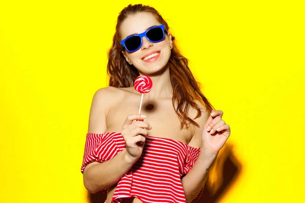 Красива молода сексуальна дівчина в блакитних сонцезахисних окулярах і червона смугаста футболка сміється і розважається і їсть цукерки на жовтому тлі — стокове фото