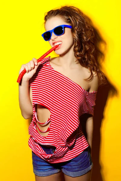 Szép fiatal szexi lány, napszemüveg kék és piros csíkos póló, nevetve és birtoklás móka, és eszik egy cukorkát, sárga háttérrel — Stock Fotó