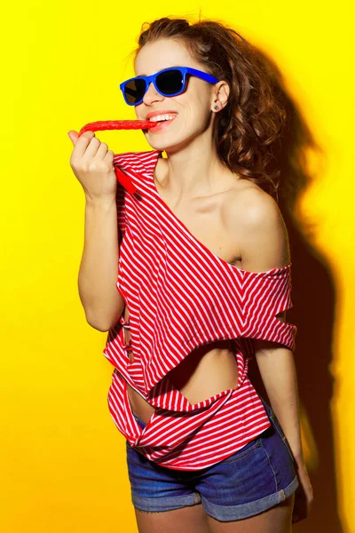 Menina sexy bonita em óculos de sol azuis e uma camiseta listrada vermelha rindo e se divertindo e comendo um doce em um fundo amarelo — Fotografia de Stock