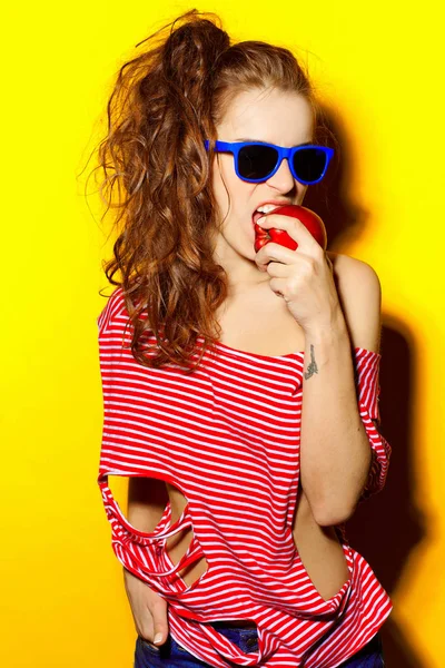 美丽年轻性感的女孩在蓝色太阳镜和红色条纹 t 恤在黄色背景上吃苹果又开心又笑 — 图库照片