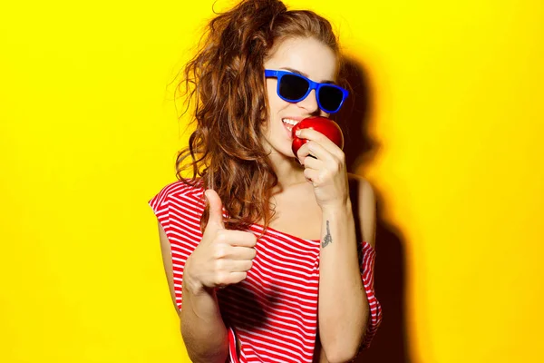 美丽年轻性感的女孩在蓝色太阳镜和红色条纹 t 恤在黄色背景上吃苹果又开心又笑 — 图库照片