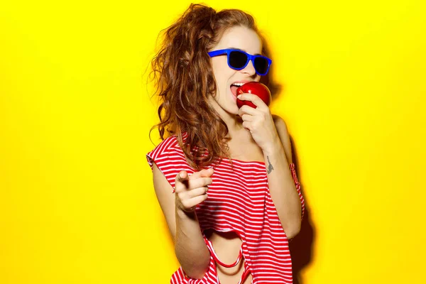 Menina sexy bonita em óculos de sol azuis e vermelho listrado T-shirt rindo e se divertindo e comendo uma maçã em um fundo amarelo — Fotografia de Stock