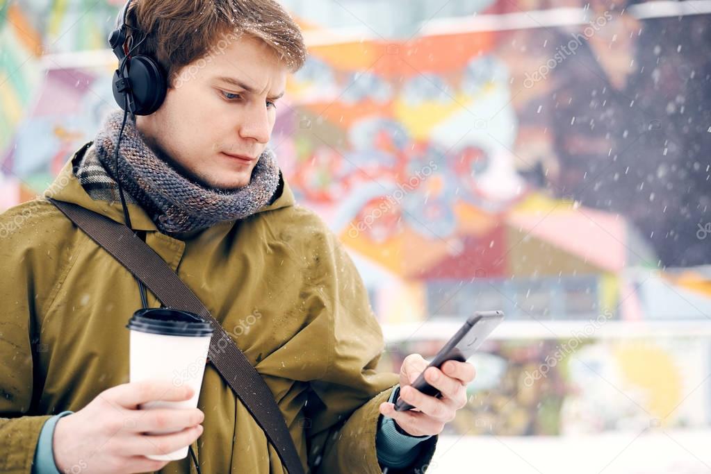 Man listening to music with headphones looking to the phone and drinking coffee in the winter on the street. Concept of technology and lifestyle