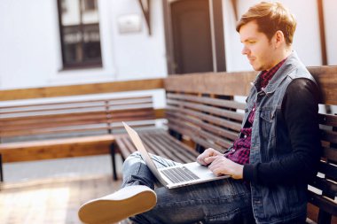 Genç adam sokakta bankta oturur ve bir dizüstü bilgisayar kullanarak. Yaşam tarzı ve teknoloji kavramı