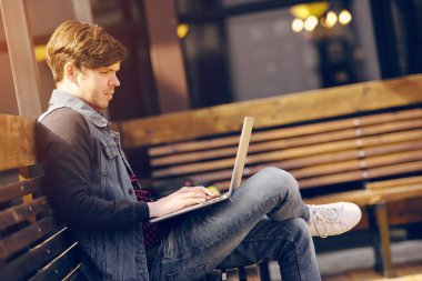 Genç adam sokakta bankta oturur ve bir dizüstü bilgisayar kullanarak. Yaşam tarzı ve teknoloji kavramı