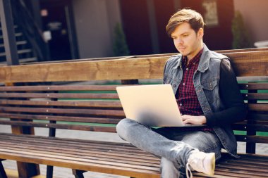 Genç adam sokakta bankta oturur ve bir dizüstü bilgisayar kullanarak. Yaşam tarzı ve teknoloji kavramı
