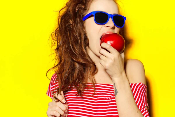 Menina sexy bonita em óculos de sol azuis e vermelho listrado T-shirt rindo e se divertindo e comendo uma maçã em um fundo amarelo — Fotografia de Stock