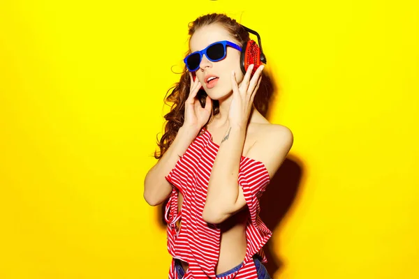 Linda menina sexy dj em óculos azuis e vermelho listrado T-shirt rindo e se divertindo e ouvindo música em fones de ouvido vermelhos em um fundo amarelo — Fotografia de Stock