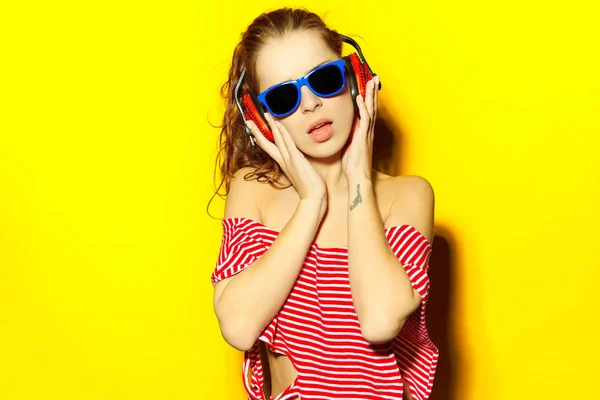 Vacker ung sexig flicka dj i blå solglasögon och röd randig T-shirt skrattar och har roligt och lyssna på musik i röda hörlurar på gul bakgrund — Stockfoto