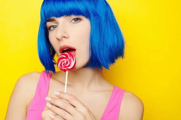在蓝色的假发和粉红色泳衣有乐趣、 在黄色背景上吃棒棒糖的性感美女 — 图库照片