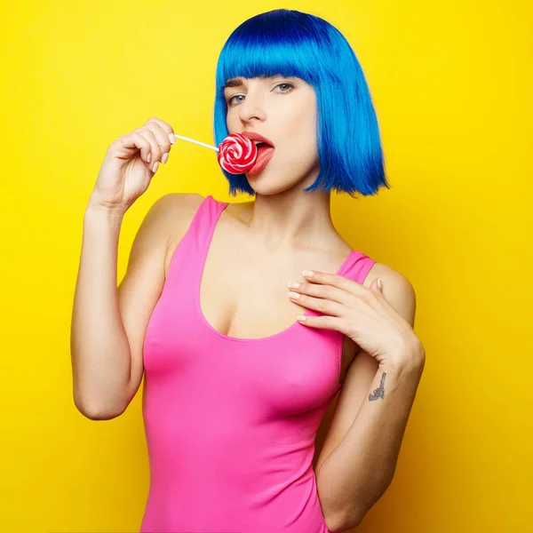 Menina sexy bonita em peruca azul e roupa de banho rosa se divertindo e comendo doces pirulito no fundo amarelo — Fotografia de Stock