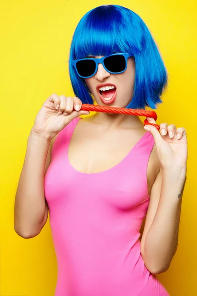 Güzel genç seksi kız mavi peruk mavi güneş gözlüğü ve pembe eğleniyor ve Sarı zemin üzerine lolipop şeker yemek mayo