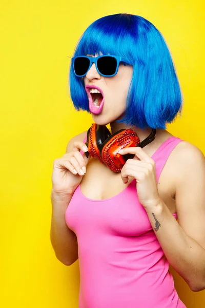 Bela jovem sexy menina dj no azul peruca azul óculos de sol e rosa maiô ter diversão e ouvir música no vermelho fones de ouvido no amarelo fundo — Fotografia de Stock