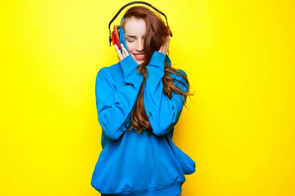 Belle jeune fille sexy DJ dans une veste bleue écoutant de la musique dans des écouteurs rouges sur un fond jaune et s'amusant — Photo